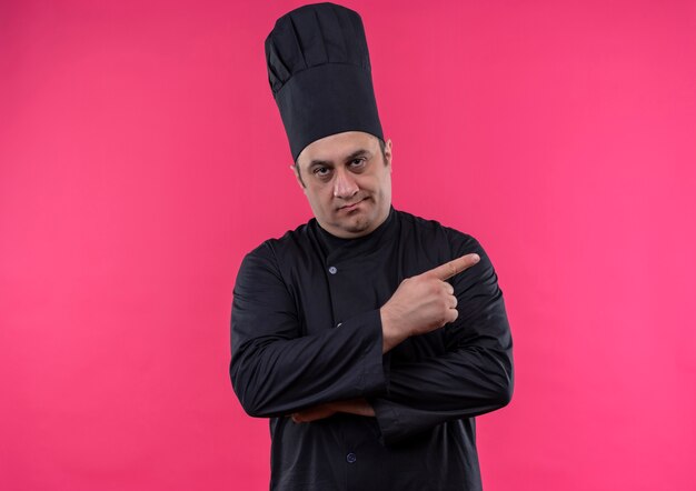 Cocinero masculino de mediana edad con uniforme de chef apunta al lado de la pared rosa aislada con espacio de copia