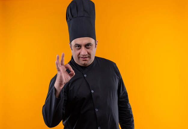 Cocinero masculino de mediana edad en uniforme de chef mostrando gesto okey en la pared amarilla