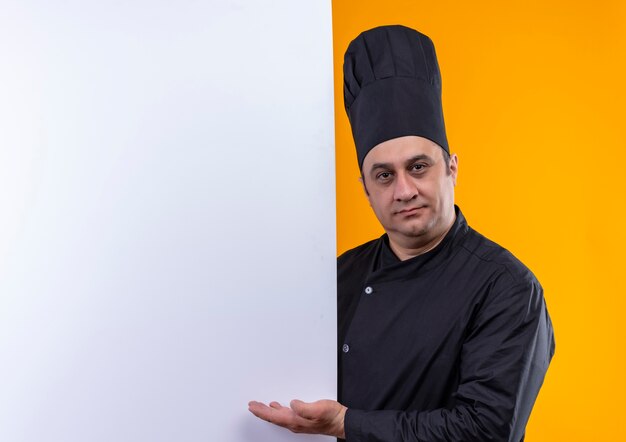 Cocinero masculino de mediana edad en uniforme de chef mostrando la pared blanca en la pared amarilla con espacio de copia