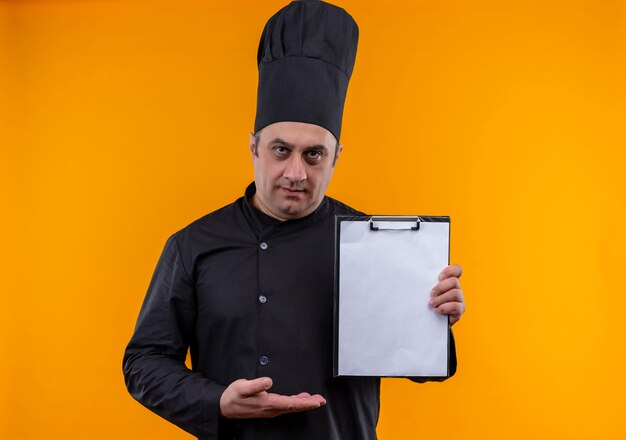 Cocinero masculino de mediana edad en uniforme de chef mostrando el portapapeles en la mano sobre la pared amarilla