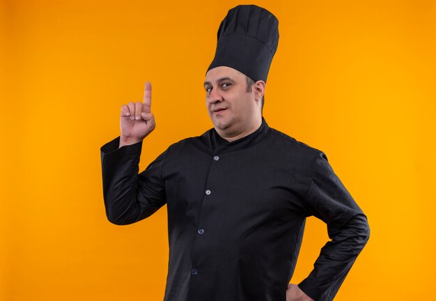 Cocinero masculino de mediana edad en uniforme de chef señala con el dedo hacia arriba poniendo la mano en la cadera en la pared amarilla con espacio de copia