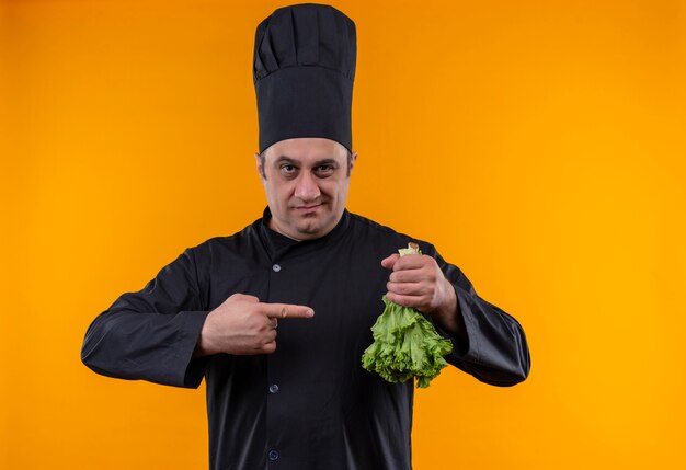 Cocinero masculino de mediana edad en uniforme de chef señala con el dedo a la ensalada en su mano sobre la pared amarilla con espacio de copia