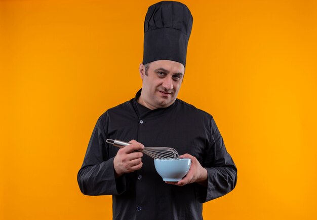 Cocinero masculino de mediana edad en uniforme de chef sosteniendo un batidor y un tazón en la pared amarilla