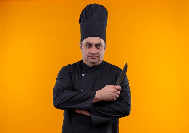 Cocinero masculino de mediana edad en uniforme de chef sosteniendo un cuchillo cruzando las manos sobre la pared amarilla con espacio de copia