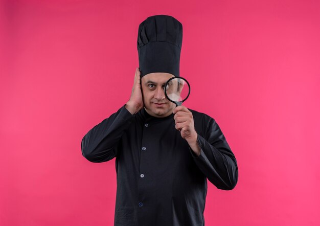 Cocinero masculino de mediana edad en uniforme de chef sosteniendo lupa poniendo la mano en la cabeza en la pared rosa aislada