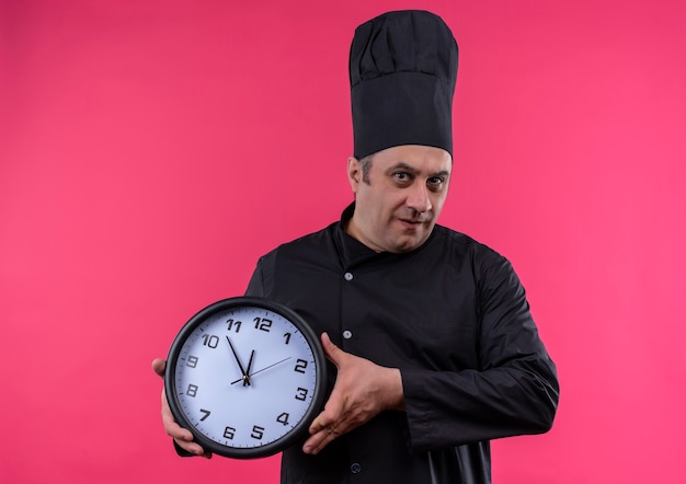 Cocinero masculino de mediana edad en uniforme de chef sosteniendo el reloj de pared en la pared rosa aislada con espacio de copia