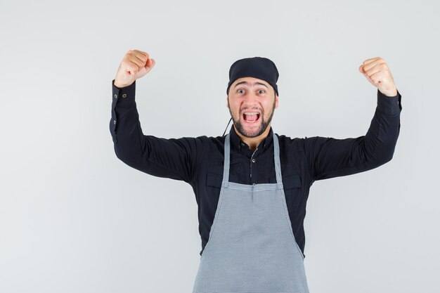 Cocinero masculino mostrando gesto de éxito en camisa, delantal y mirando dichoso, vista frontal.