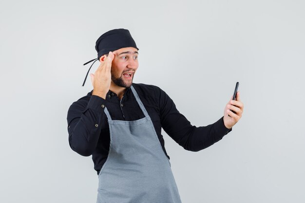Cocinero masculino posando mientras toma selfie en camisa, delantal y mirando alegre. vista frontal.