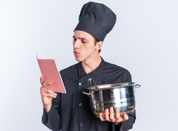 Cocinero masculino rubio joven concentrado en uniforme del cocinero y la tapa que sostiene el bloc de notas de lectura de la olla