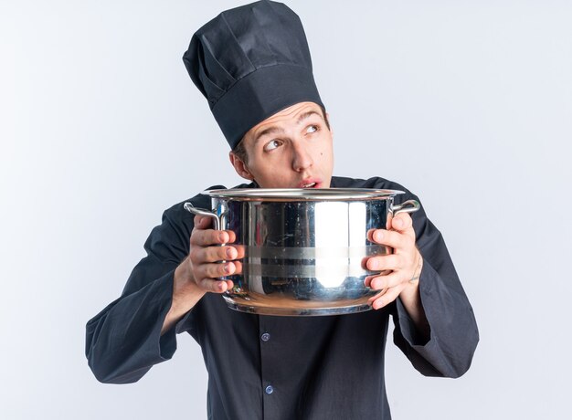 Cocinero masculino rubio joven pensativo en uniforme del cocinero y la tapa que sostiene la olla mirando hacia arriba
