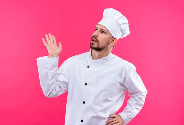 Cocinero profesional masculino en uniforme blanco y sombrero de cocinero mirando hacia arriba con expresión confusa sobre fondo rosa