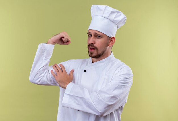 Cocinero profesional de sexo masculino en uniforme blanco y gorro de cocinero mostrando bíceps mirando satisfecho y orgulloso de pie sobre fondo verde