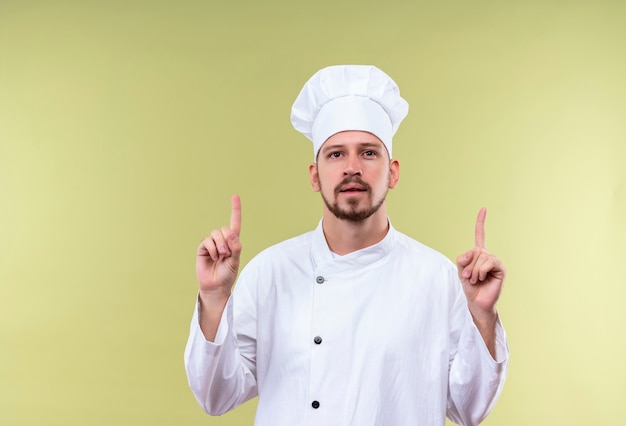 Cocinero profesional de sexo masculino en uniforme blanco y sombrero de cocinero apuntando hacia arriba con los dedos teniendo una nueva idea de pie sobre fondo verde