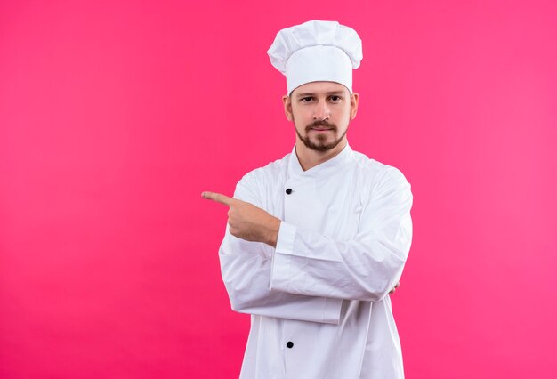 Cocinero profesional de sexo masculino en uniforme blanco y sombrero de cocinero apuntando con el dedo hacia el lado mirando a la cámara con expresión segura de pie sobre fondo rosa