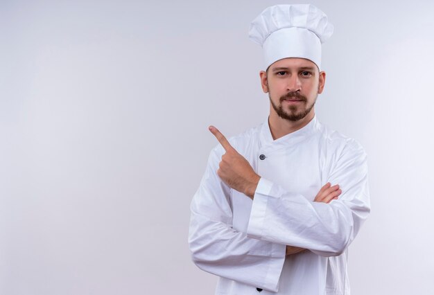Cocinero profesional de sexo masculino en uniforme blanco y sombrero de cocinero apuntando hacia el lado con el dedo índice mirando confiado de pie sobre fondo blanco