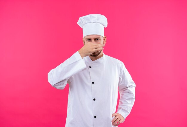 Cocinero profesional de sexo masculino en uniforme blanco y sombrero de cocinero cerrando la nariz, concepto de mal olor sobre fondo rosa