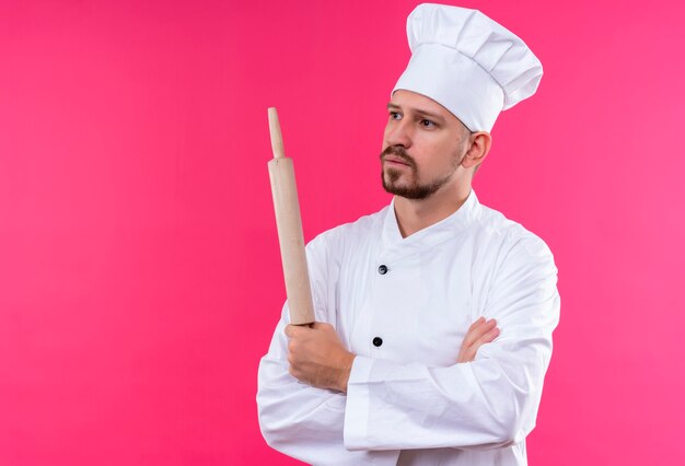 Cocinero profesional de sexo masculino en uniforme blanco y sombrero de cocinero de pie con los brazos cruzados sosteniendo el rodillo con expresión pensativa en la cara sobre fondo rosa