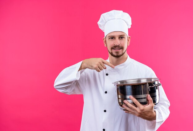 Cocinero profesional de sexo masculino en uniforme blanco y sombrero de cocinero sosteniendo una cacerola vacía apuntando con el dedo a ella sonriendo confiada de pie sobre fondo rosa