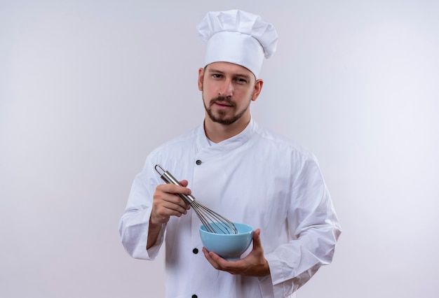 Cocinero profesional de sexo masculino en uniforme blanco y sombrero de cocinero sosteniendo un cuenco batiendo algo con un batidor mirando confiado de pie sobre fondo blanco.