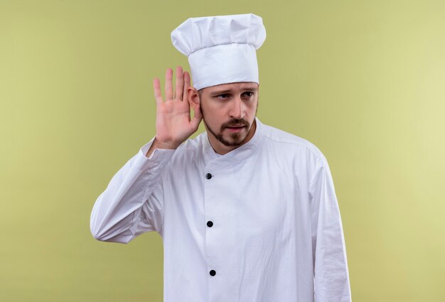 Cocinero profesional de sexo masculino con uniforme blanco y sombrero de cocinero sosteniendo la mano cerca de la oreja tratando de escuchar la conversación de alguien de pie sobre fondo verde