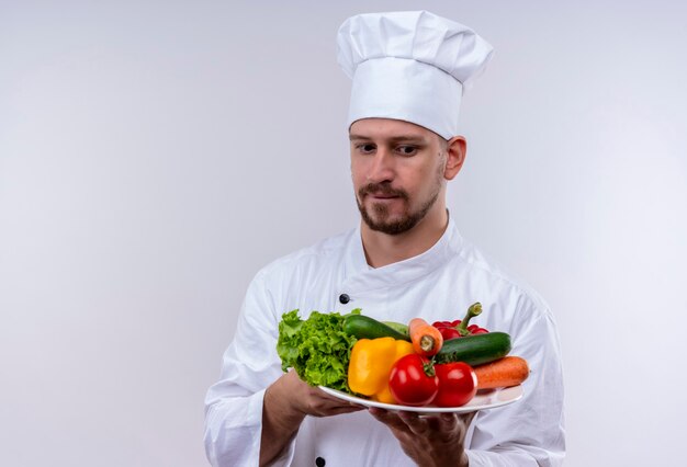 Cocinero profesional de sexo masculino en uniforme blanco y sombrero de cocinero sosteniendo un plato con verduras mirándolos intrigado de pie sobre fondo blanco