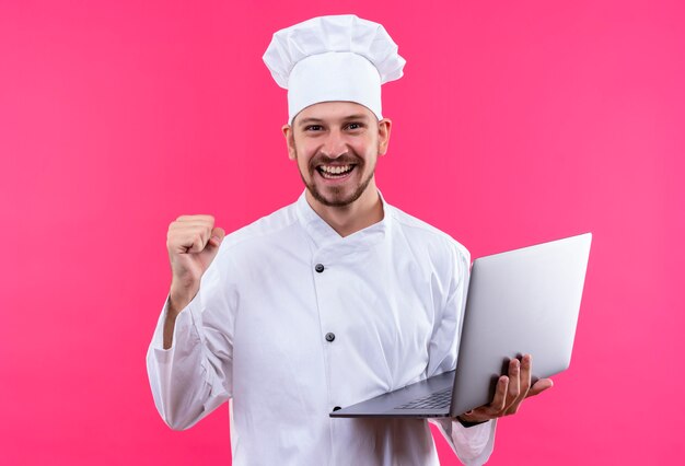 Cocinero profesional de sexo masculino en uniforme blanco y sombrero de cocinero sosteniendo portátil mirando a cámara sonriendo alegremente levantando el puño regocijándose de su éxito de pie sobre fondo rosa