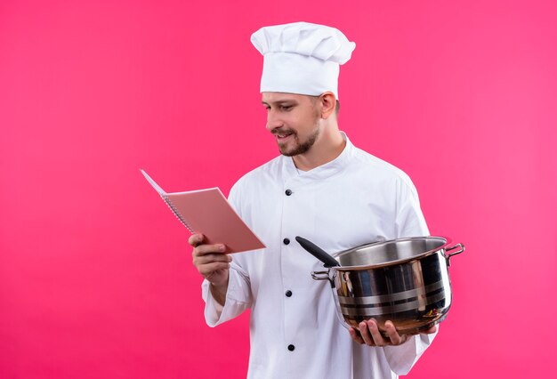 Cocinero profesional de sexo masculino en uniforme blanco y sombrero de cocinero sosteniendo una sartén y un cuaderno mirándolo sonriendo de pie sobre fondo rosa