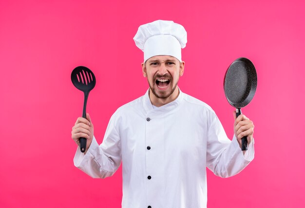 Cocinero profesional de sexo masculino en uniforme blanco y sombrero de cocinero sosteniendo una sartén y un cucharón gritando con expresión agresiva, frustrado de pie sobre fondo rosa