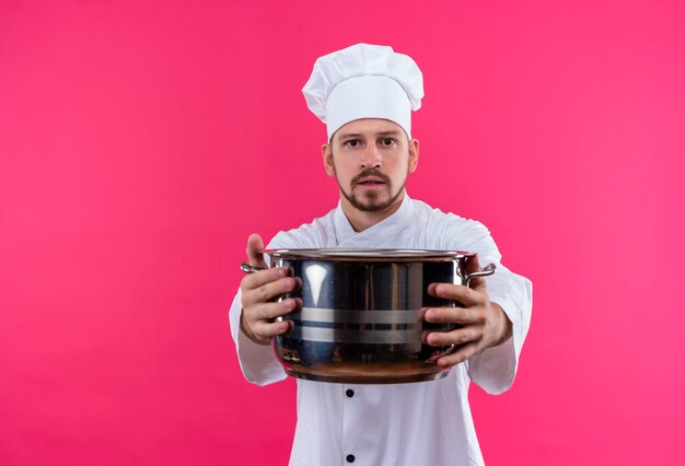 Cocinero profesional de sexo masculino en uniforme blanco y sombrero de cocinero sosteniendo una sartén vacía mirando a la cámara con cara seria sobre fondo rosa