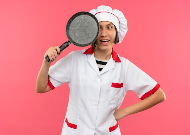 Cocinero de sexo femenino joven alegre en uniforme del cocinero que sostiene y que mira la sartén aislada en rosa