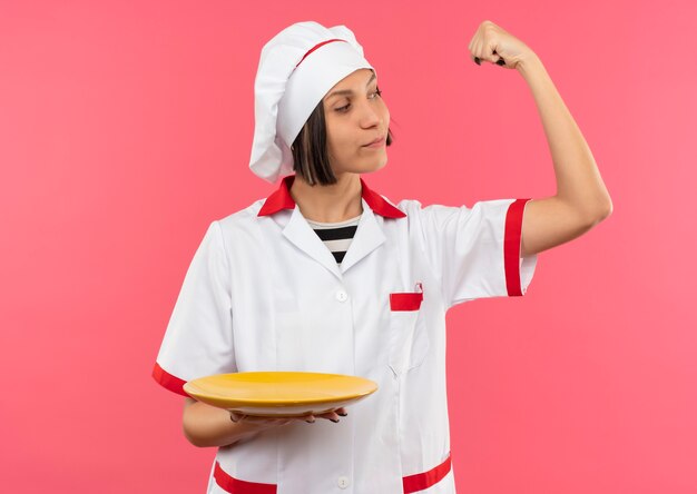 Cocinero de sexo femenino joven confiado en uniforme del cocinero que gesticula el plato fuerte que sostiene y que mira su brazo aislado en rosa