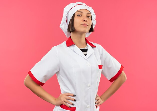 Foto gratuita cocinero de sexo femenino joven confiado en uniforme del cocinero que pone las manos en la cintura aislada en rosa