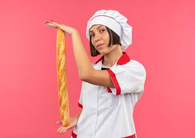 Cocinero de sexo femenino joven confiado en uniforme del cocinero que sostiene el palillo de pan que mira aislado en rosa