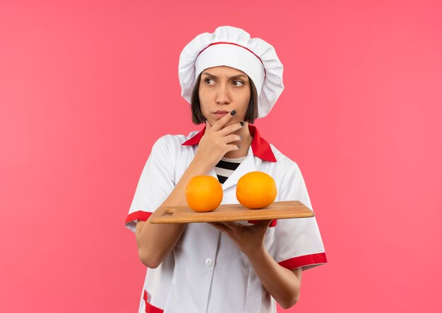 Cocinero de sexo femenino joven pensativo en uniforme del cocinero que sostiene la tabla de cortar con las naranjas en él que toca su barbilla y que mira el lado aislado en la pared rosada