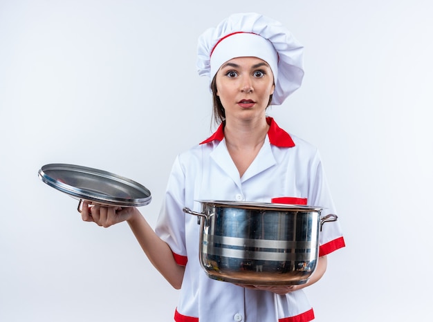 Cocinero de sexo femenino joven sorprendido vistiendo uniforme del cocinero que sostiene la cacerola con la tapa aislada en el fondo blanco