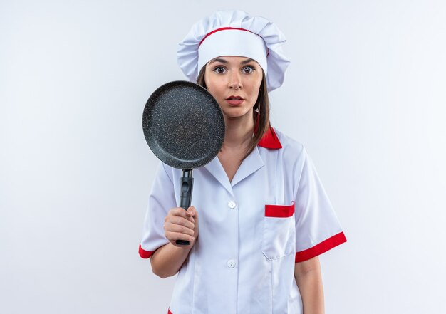 Cocinero de sexo femenino joven sorprendido vistiendo uniforme del cocinero que sostiene la sartén aislada en el fondo blanco