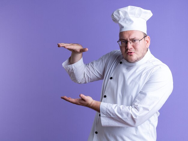 Cocinero de sexo masculino adulto con el ceño fruncido vistiendo uniforme de chef y gafas de pie en la vista de perfil mirando al frente mostrando gesto de tamaño aislado en la pared púrpura con espacio de copia