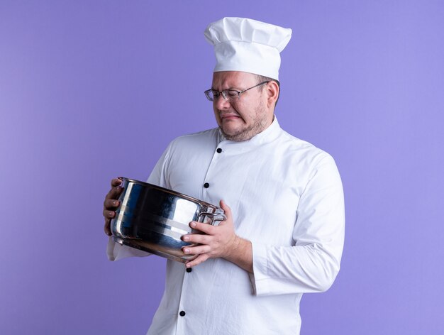 Cocinero de sexo masculino adulto con el ceño fruncido vistiendo uniforme de chef y gafas sosteniendo y mirando dentro de la olla aislado en la pared púrpura