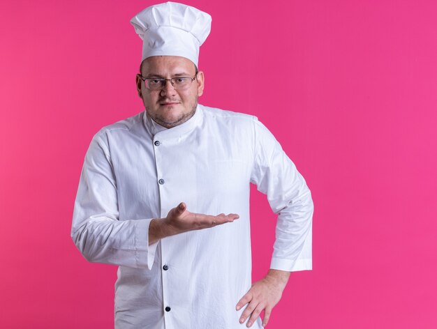 Cocinero de sexo masculino adulto complacido vistiendo uniforme de chef y gafas manteniendo la mano en la cintura mostrando la mano vacía mirando al frente aislado en la pared rosa con espacio de copia