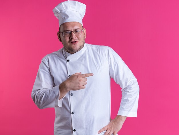 Cocinero de sexo masculino adulto complacido vistiendo uniforme de chef y gafas mirando al frente manteniendo la mano en la cintura apuntando al lado aislado en la pared rosa