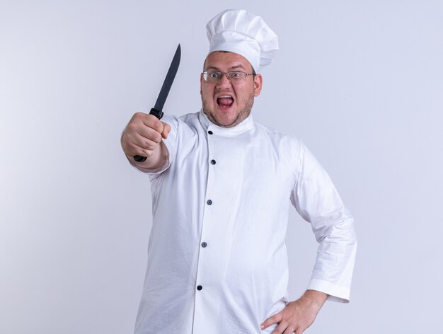 Cocinero de sexo masculino adulto furioso con uniforme de chef y gafas manteniendo la mano en la cintura mirando al frente estirando el cuchillo hacia el frente aislado en la pared blanca