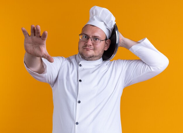 Cocinero de sexo masculino adulto impresionado con uniforme de chef y gafas mirando a la cámara sosteniendo la tapa de la olla detrás de la cabeza tocando la cabeza con ella estirando la mano hacia la cámara aislada sobre fondo naranja