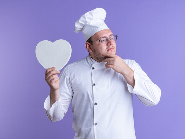 Cocinero de sexo masculino adulto reflexivo que lleva el uniforme del cocinero y los vidrios aislados en mantener la mano en la barbilla sosteniendo la forma del corazón que mira el lado de la pared púrpura