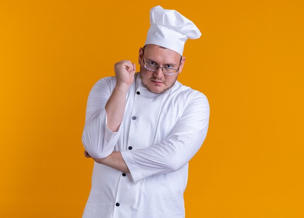 Cocinero de sexo masculino adulto seguro con uniforme de chef y gafas mirando al frente manteniendo la mano debajo del codo y el puño en el aire aislado en la pared naranja con espacio de copia