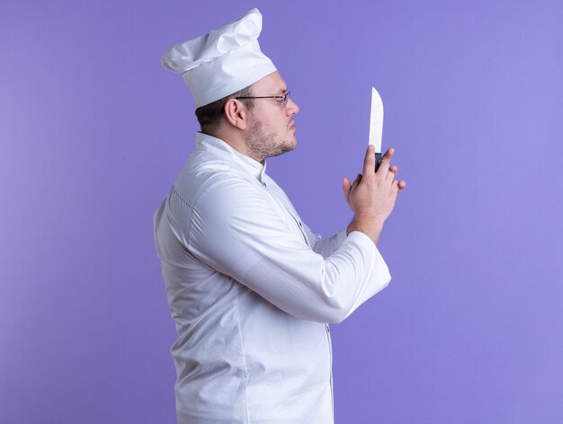 Cocinero de sexo masculino adulto serio vistiendo uniforme de chef y gafas de pie en la vista de perfil sosteniendo y mirando el cuchillo aislado en la pared púrpura con espacio de copia