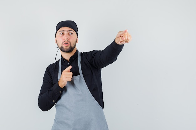 Cocinero de sexo masculino apuntando hacia afuera en camisa, delantal y mirando enfocado. vista frontal.