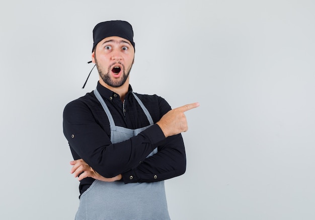 Cocinero de sexo masculino apuntando hacia afuera en camisa, delantal y mirando sorprendido. vista frontal.