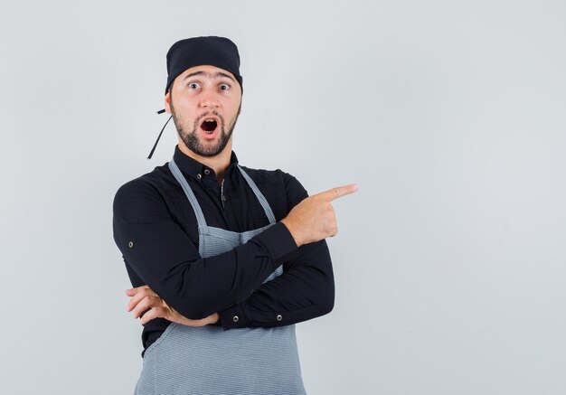 Cocinero de sexo masculino apuntando hacia afuera en camisa, delantal y mirando sorprendido. vista frontal.