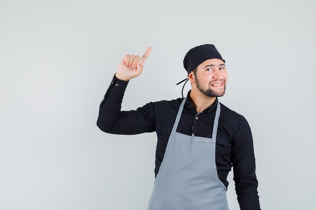 Foto gratuita cocinero de sexo masculino apuntando hacia arriba en camisa, delantal y mirando alegre. vista frontal.