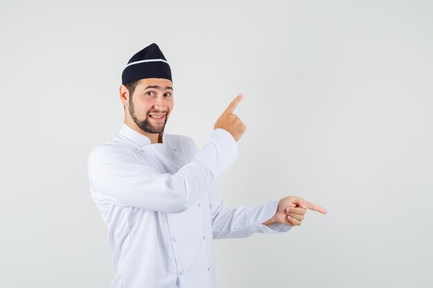 Cocinero de sexo masculino apuntando a un lado en uniforme blanco y mirando alegre. vista frontal.